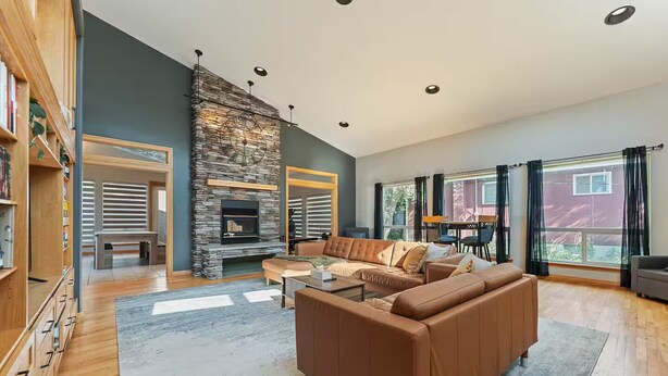 A spacious living room with a stone fireplace, leather sofa, and large windows offering a view. - Riverfront City Escape w/Hot Tub, 2 King, Gym, Sleeps 12, Firepit, Kayaks (Winnipeg)