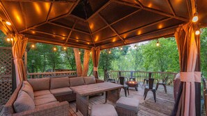 An outdoor deck with a wooden bench, a sofa, and chairs, surrounded by trees. - Riverfront City Escape w/Hot Tub, 2 King, Gym, Sleeps 12, Firepit, Kayaks (Winnipeg)