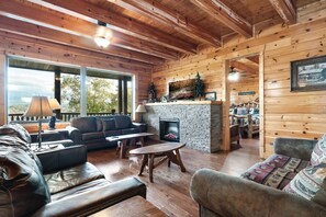 L’intérieur d’une cabane en rondins, avec une cheminée, des meubles en bois et une grande fenêtre donnant sur un paysage.