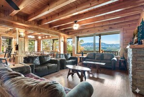 L’intérieur d’une cabane en rondins, avec une cheminée, des meubles en bois et de grandes fenêtres donnant sur les montagnes.