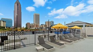 A rooftop pool area with lounge chairs and umbrellas, surrounded by a metal fence, overlooking a city skyline with tall buildings. - King Bed Suite | Rooftop Pool | Gym | Private Balcony | Midtown ATL (Atlanta)