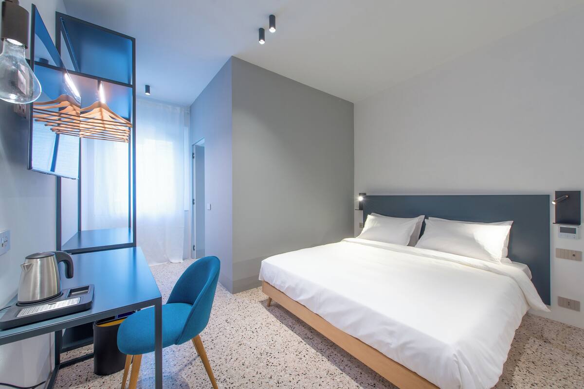 Ein modernes Hotelzimmer mit einem Bett, einem Schreibtisch mit Wasserkocher, einem Sessel und einem Spiegel.