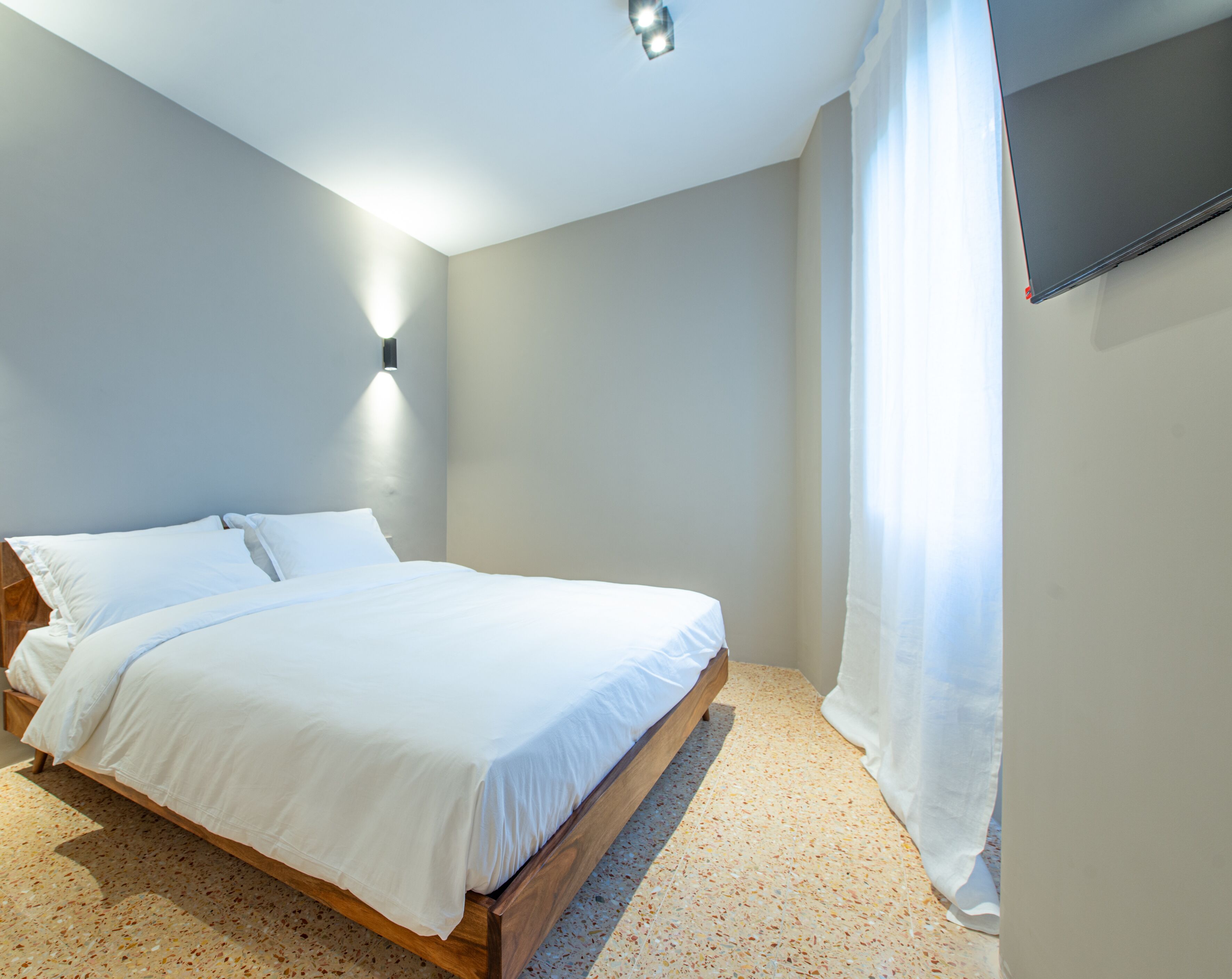 A bedroom with a wooden bed, white bedding, a wall-mounted TV, and a ceiling light.