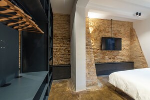 Una camera da letto con soffitto spiovente, pareti in mattoni a vista, una TV a schermo piatto montata al muro e un letto con copripiumino bianco.
