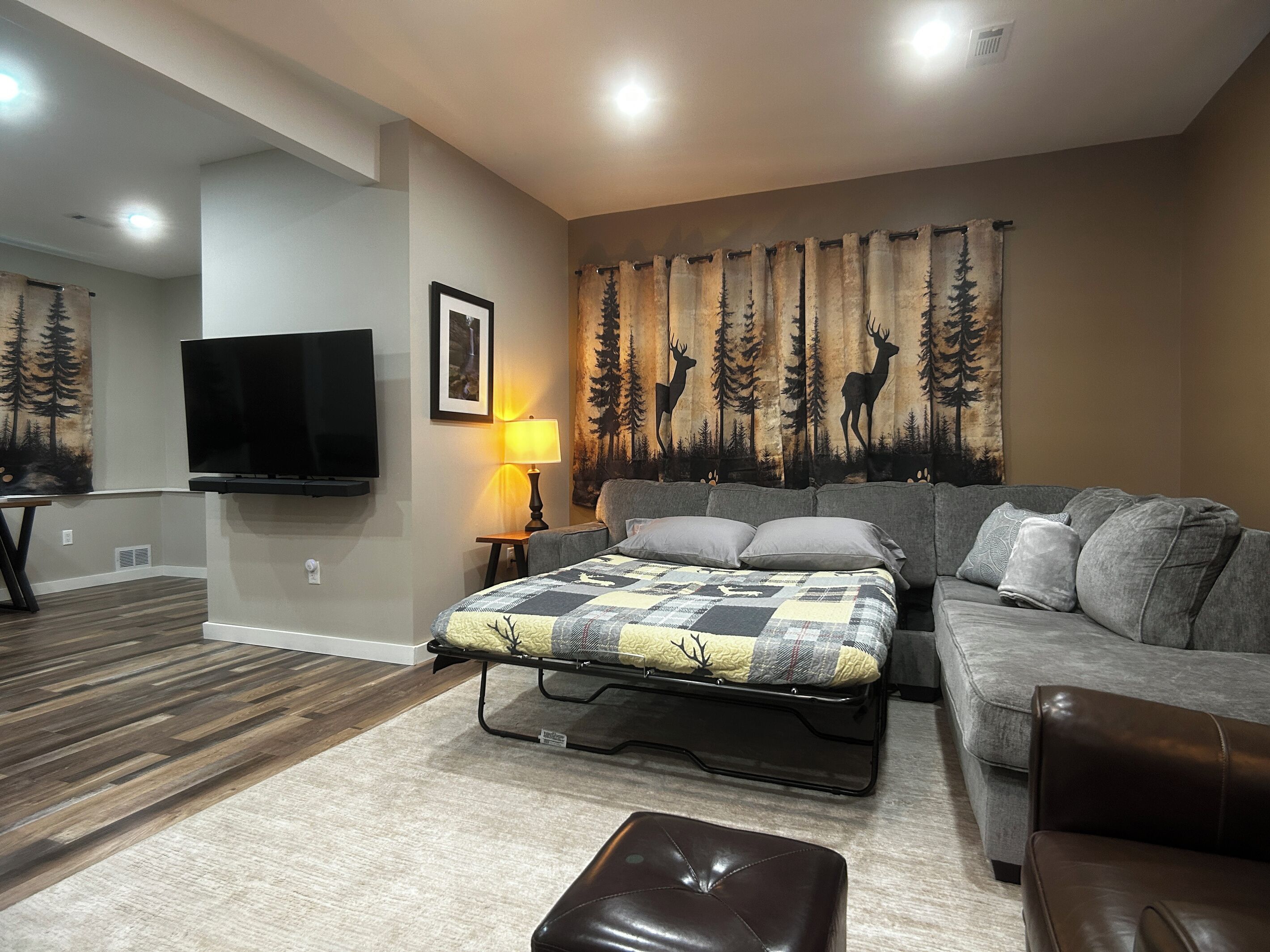 A bedroom with a bed, a grey sofa, a brown leather ottoman, a TV, a lamp, and a forest-themed curtain.