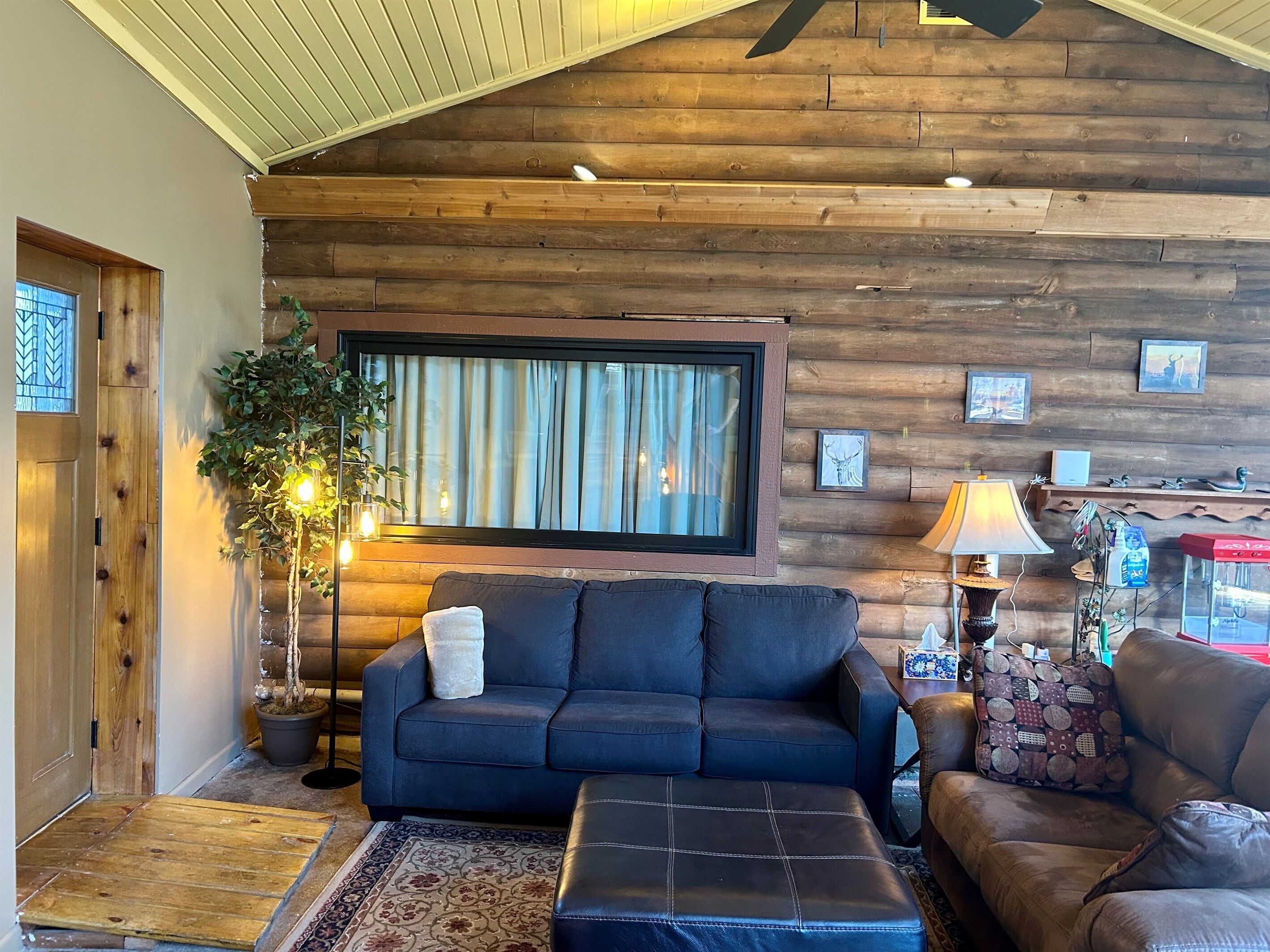 A cozy living room with a wooden wall, a large window, a sofa, a coffee table, and a lamp.