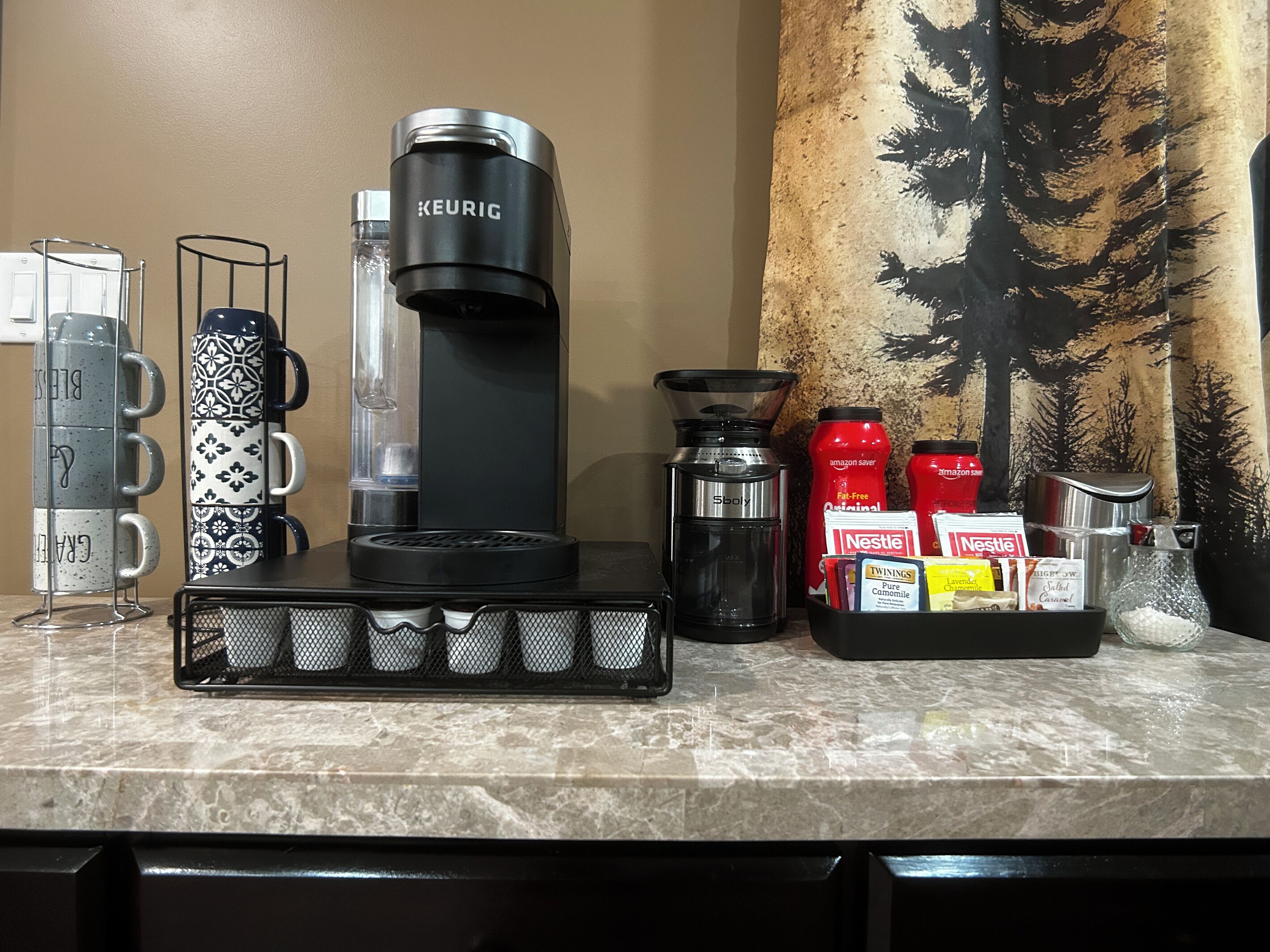 Eine Küchenarbeitsplatte mit einer Keurig-Kaffeemaschine, einem Spoliy-Wasserspender und einer Kaffeemaschine. Außerdem liegen zwei Nestle-Kaffeekapseln und eine rote Amazon Saver Original-Flasche bereit.