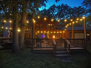 A wooden cabin with a deck, string lights, and a fire.