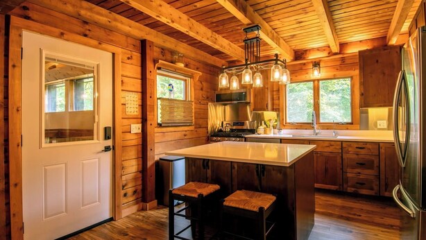 A wooden cabin kitchen with a central island, bar stools, and a hanging light fixture.