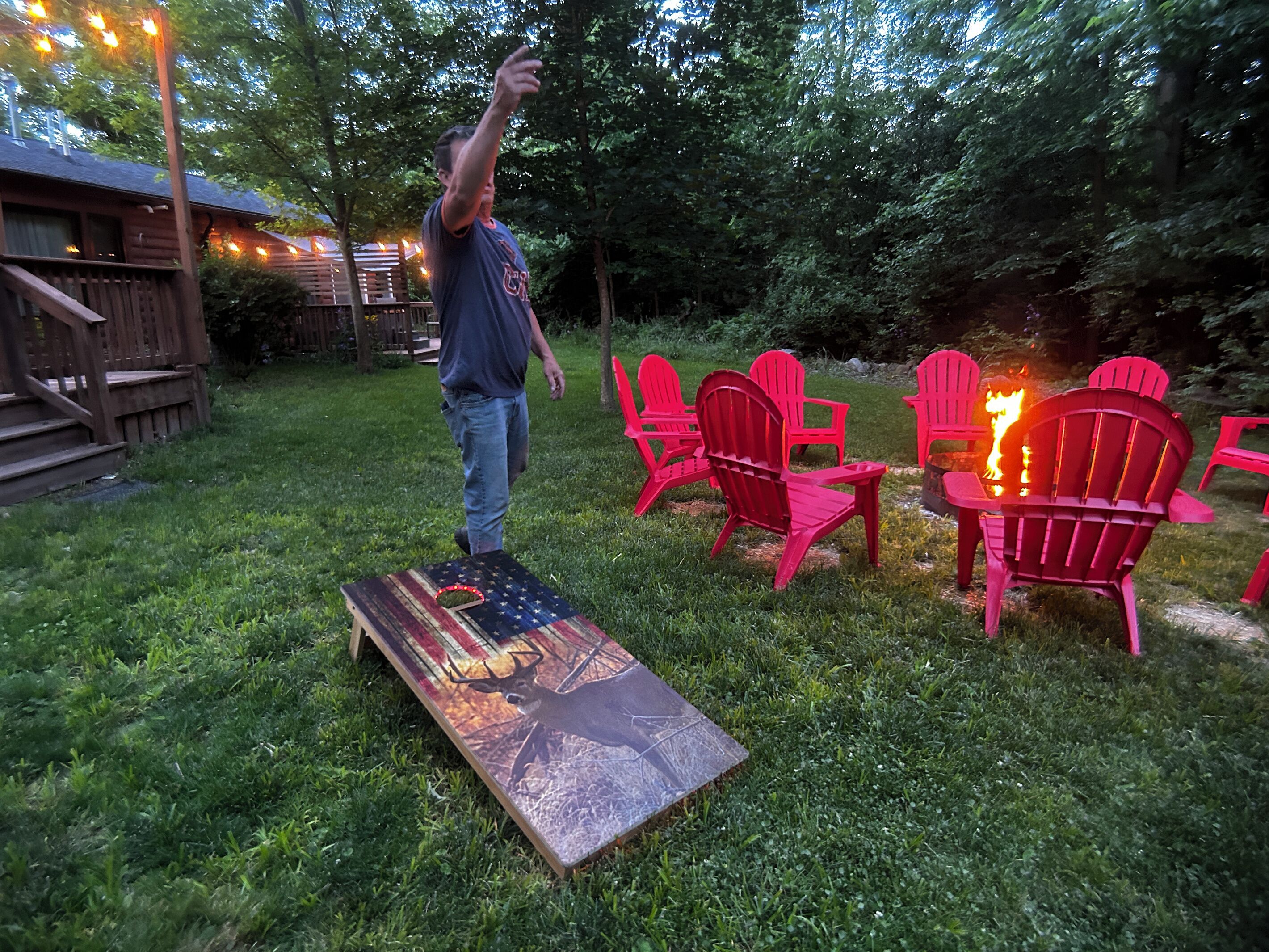Une personne se tient dans un espace extérieur verdoyant, avec une terrasse en bois, des chaises Adirondack rouges et un foyer.
