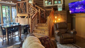 A cozy cabin interior with a wooden staircase, a dining area with a glass table, and a leather armchair.