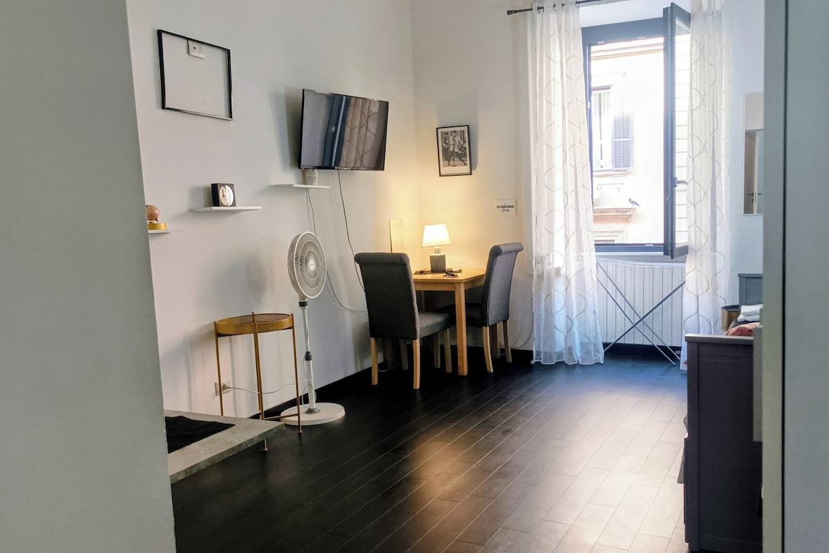 Una habitación pequeña y bien iluminada, con una mesa de comedor y sillas, un ventilador de pie, un televisor montado en la pared y una ventana con cortinas.