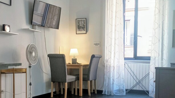 A modern living room with a flat-screen TV, a standing fan, a dining table with chairs, and a large window with curtains. - Little Appartment (Rome)