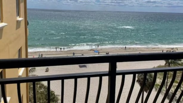 Vista del océano desde un balcón con barandilla, una playa de arena y cielo despejado.
