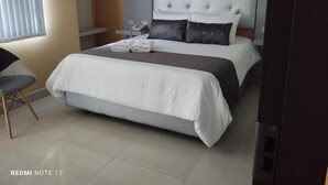 Ein ordentlich bezogenes Bett mit grauem Kopfteil, ein Nachttisch mit Lampe und ein Stuhl.