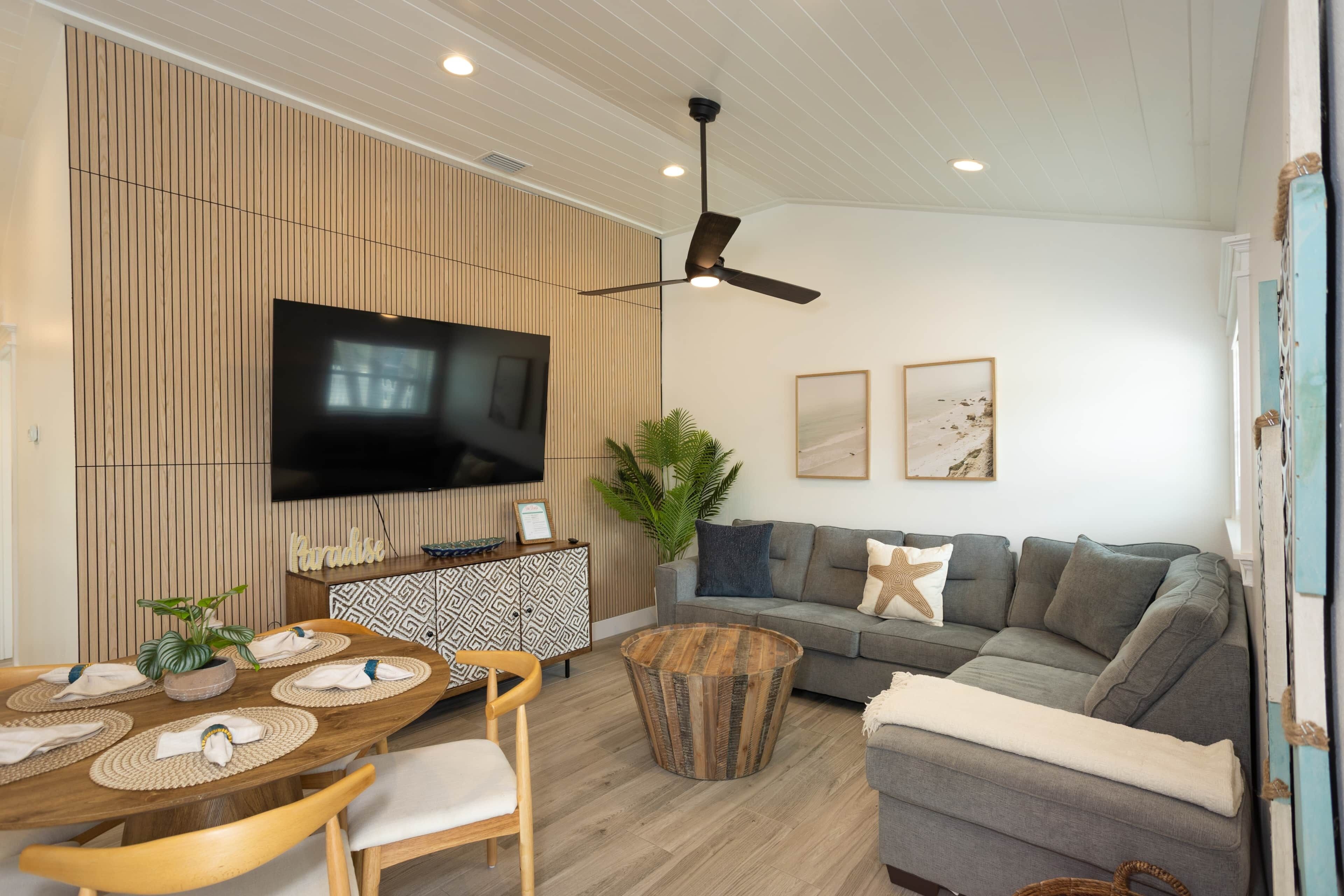 Un salon moderne doté d’un téléviseur à écran plat, d’un ventilateur de plafond, d’un canapé gris et d’une table à manger en bois avec des sets de table.