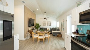 A modern kitchen with a built-in oven, a dining area with a round table and chairs, a grey sofa, and a ceiling fan.