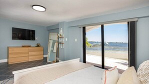 A bedroom with a large bed, a wooden dresser, a TV on the wall, and a view of the beach through a sliding door. - Waterfront 2BR on Manatee River Near Anna Maria Island (West Bradenton)