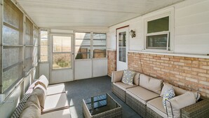 Un patio couvert comprenant un canapé en osier, une table basse en verre et une porte blanche donnant sur l’extérieur.