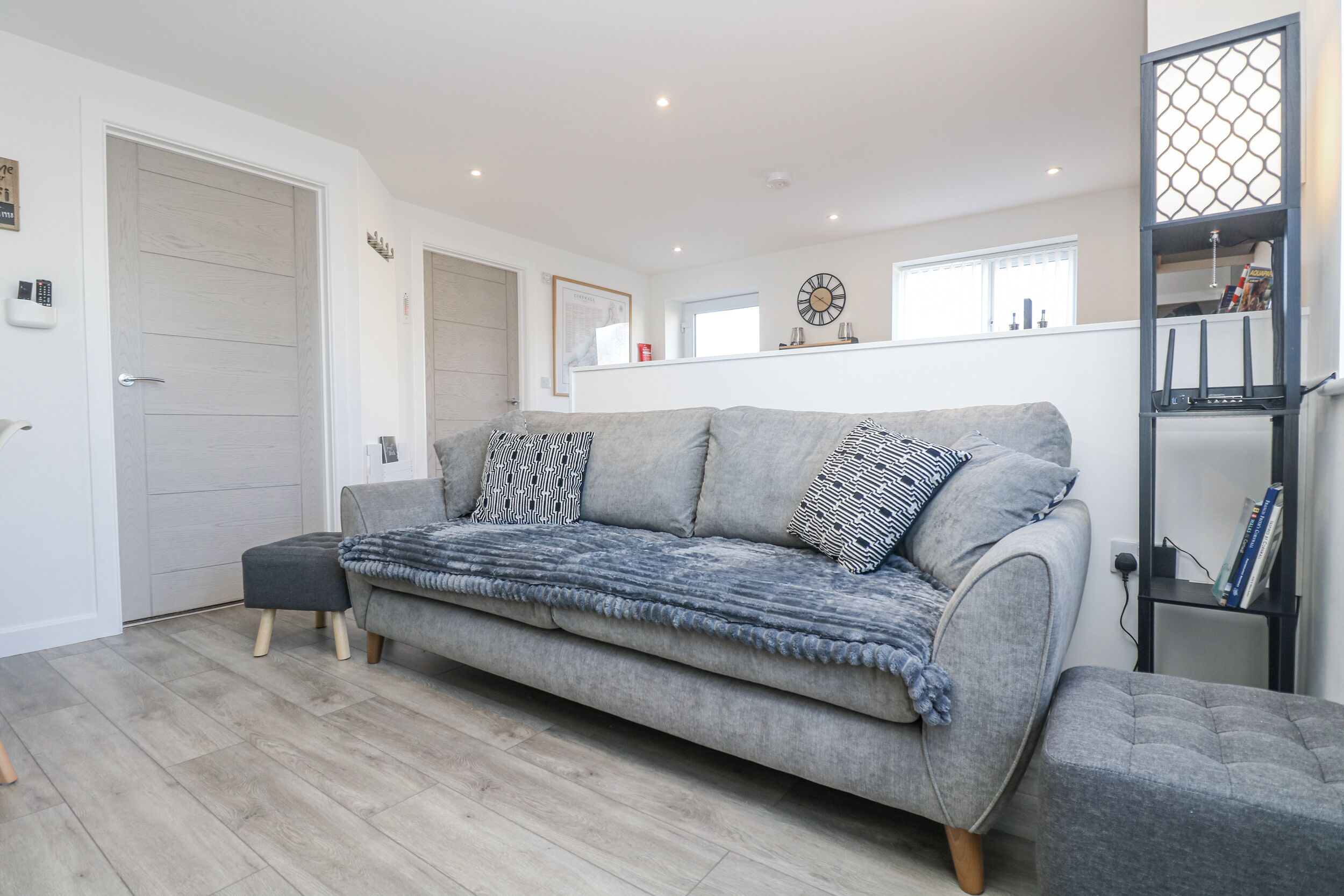 A modern living room with a grey sofa, patterned cushions, a wooden floor, a bookshelf, and a wall-mounted clock.