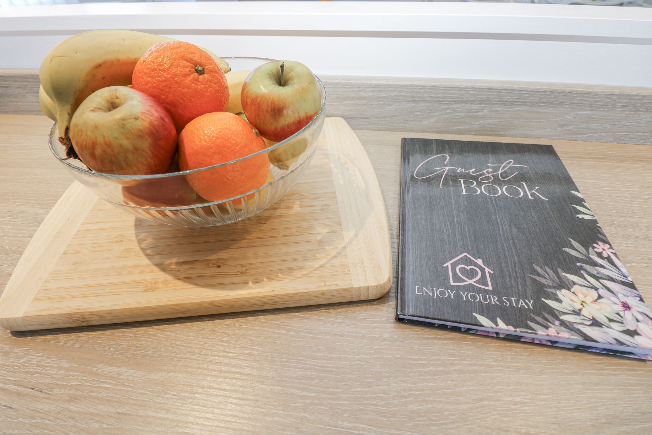 A bowl of fruit on a wooden board next to a book titled "Guest Book" with the subtitle "Enjoy Your Stay".