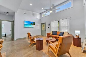A spacious living room with a sofa, armchairs, and a coffee table. A television is mounted on the wall, and there is a door leading to another room.