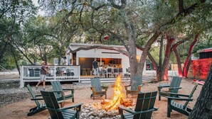 An outdoor seating area with chairs arranged around a fire pit, a building with a covered patio, and trees in the background.