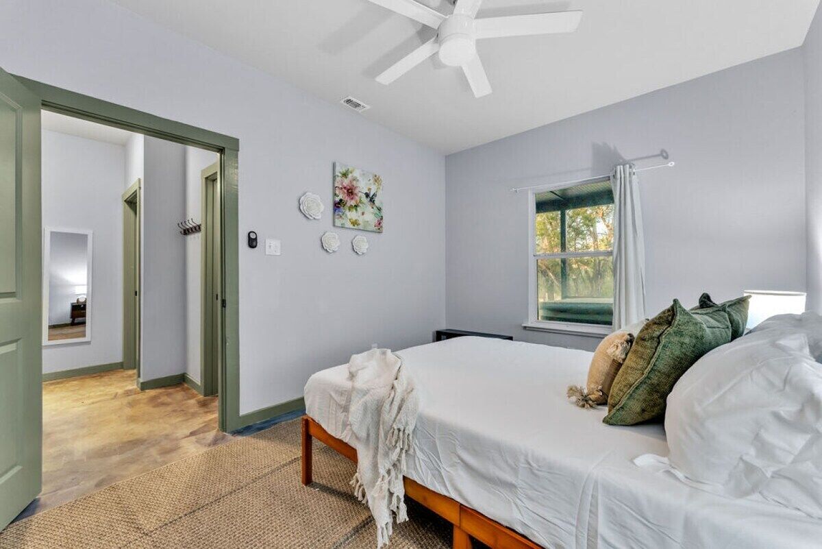 Een slaapkamer met een bed, een raam met gordijnen, een plafondventilator en een deur naar een andere kamer.