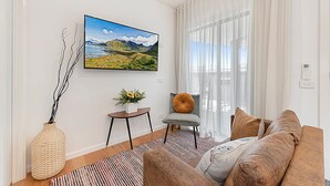 A modern living room with a large flat-screen TV mounted on the wall, a comfortable sofa, a small side table with a vase of flowers, and a view of a mountain landscape through sheer curtains. - Gorgeous 3-bed House With Spacious Shared Backyard (Melbourne)