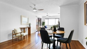 A spacious room with a wooden floor, a round dining table with chairs, a ceiling fan, and a large window providing natural light. - Coastal 2-bed With Balcony, 200m From Beach (Gold Coast)