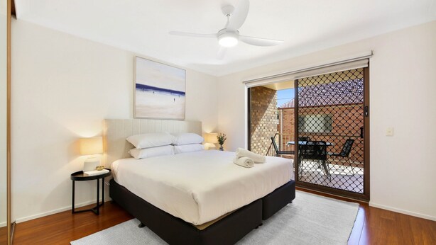 A bedroom with a bed, bedside lamps, a ceiling fan, a view through a sliding door to an outdoor area with a table and chairs. - Coastal 2-bed With Balcony, 200m From Beach (Gold Coast)