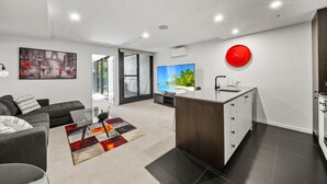 A modern living room with a sofa, a coffee table, and a TV. There's a kitchen area with a sink and cabinets, and a red clock on the wall. - Central 1-bed Apartment With Pool (Canberra)