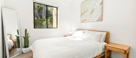 A bedroom with a wooden bed, white bedding, a nightstand, a mirror, a cactus plant, and a window with a view of greenery.