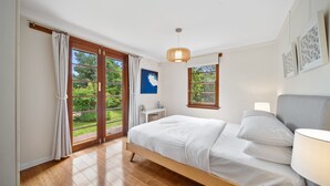 Een slaapkamer met een houten bed, een nachtkastje, een lamp en uitzicht op groen door grote schuifdeuren.