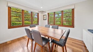Een ruime eethoek met een houten tafel en stoelen, grote ramen met uitzicht op groen, en een wit plafond met inbouwverlichting.