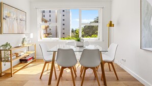 Een moderne eetruimte met een glazen tafel, witte stoelen en uitzicht op gebouwen door grote ramen.
