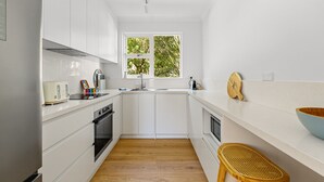 Een moderne keuken met witte kasten, een houten vloer, een raam met uitzicht op bomen, een houten kom op het aanrecht en een broodrooster.