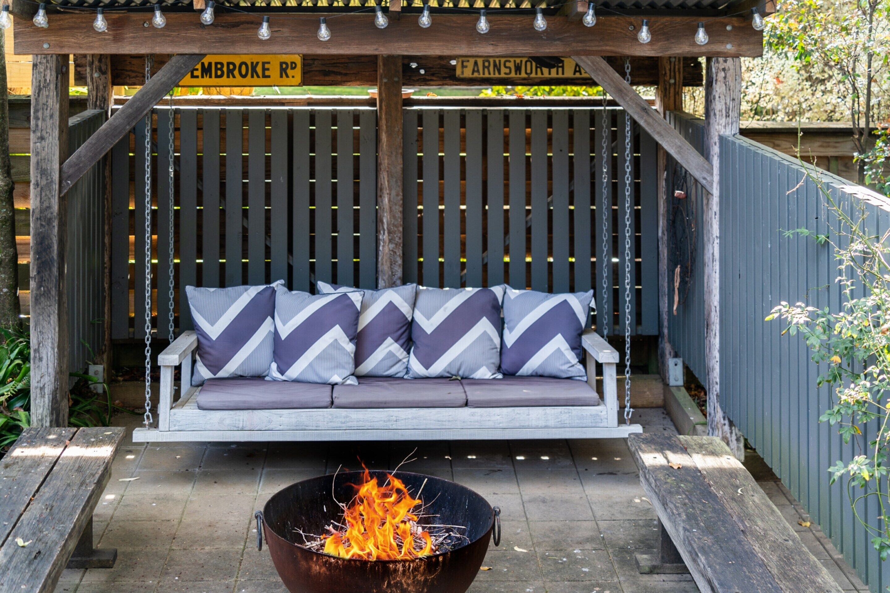A wooden swing seat with chevron-patterned cushions, a fire pit in the foreground, and a wooden pergola overhead.