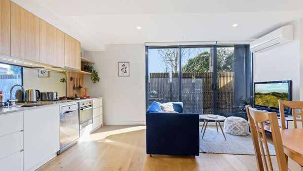 A modern kitchen with white cabinets, stainless steel appliances, and a dining area with a dark blue sofa and wooden table. - Cosy 1-bed With Private Balcony By Showgrounds (Melbourne)