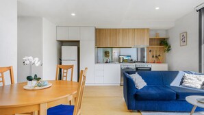 A modern kitchen with wooden cabinets, a dining table with blue chairs, and a blue sofa in the living area. - Cosy 1-bed With Private Balcony By Showgrounds (Melbourne)