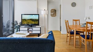A modern living room with a flat-screen TV, a sofa, a dining table with chairs, and a large window with plants. - Cosy 1-bed With Private Balcony By Showgrounds (Melbourne)