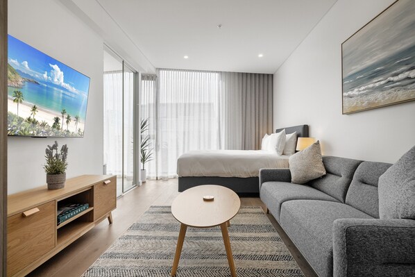 Una habitación de hotel moderna con una cama, un sofá gris, una mesita de madera y un televisor grande en la pared.