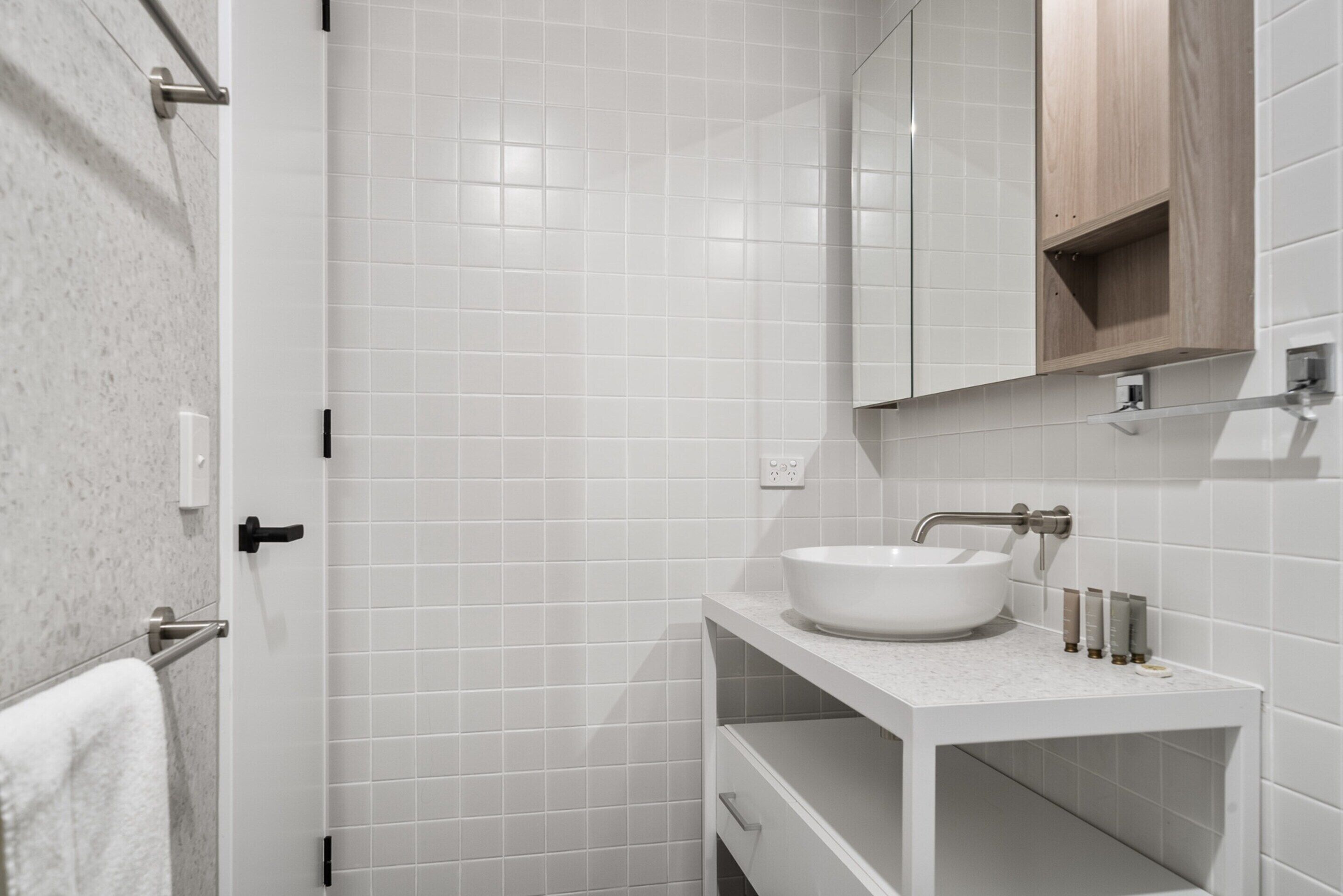 A modern bathroom with white tiles, a white sink, and a wooden cabinet above the mirror.