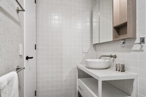 Un baño moderno con azulejos blancos, un lavabo blanco y un mueble de madera sobre el espejo.