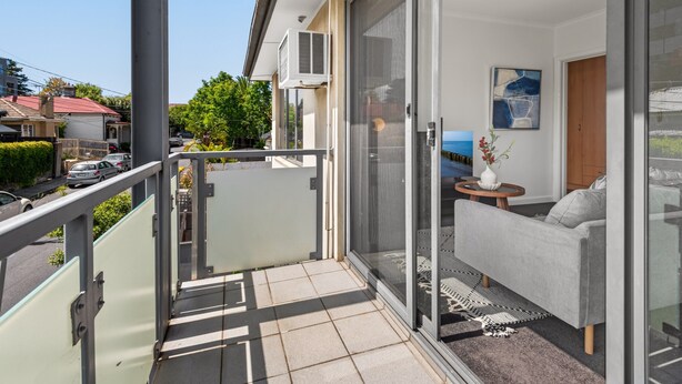 Un balcon avec une porte-fenêtre coulissante, un canapé gris, une petite table ronde et un vase rempli de fleurs.