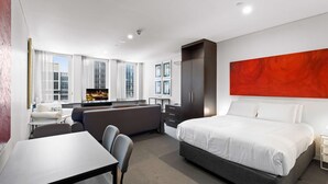 A modern hotel room with a large bed, a desk, a sofa, and a view of the cityscape through the windows. - Cosy Studio in the City Centre (Melbourne)