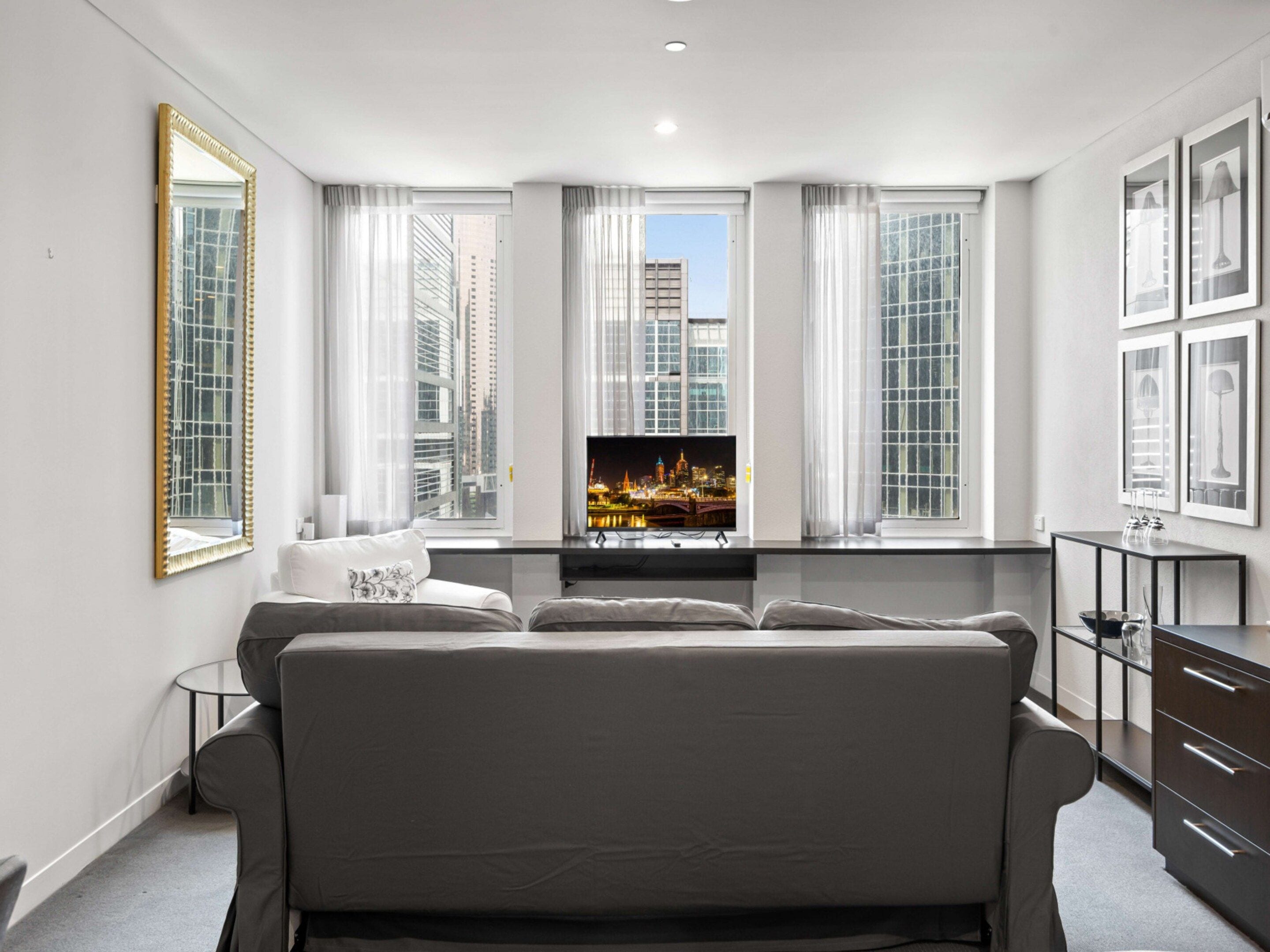 A modern living room with a sofa, a TV, and a cityscape view through large windows.