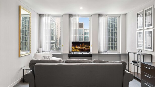 A modern living room with a sofa, a flat-screen TV, and a city view through large windows. - Cosy Studio in the City Centre (Melbourne)