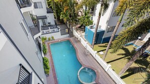 Une piscine sur le toit, dotée d’un jacuzzi circulaire, entourée d’une clôture blanche, de palmiers et offrant une vue sur les bâtiments adjacents.