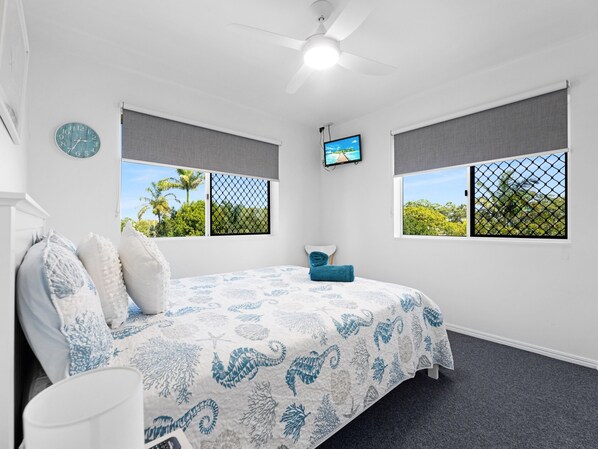 A bedroom with a bed, two windows with view screens, a ceiling fan, a clock, and a TV on the wall. - 2-bed Escape With Pool, Steps From Torquay Beach (Hervey Bay)
